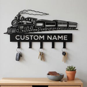May include: Black metal key holder featuring a train design with the text "CUSTOM NAME". The key holder has four hooks and is mounted on a white wall. Keys hang from the hooks. A wooden cabinet with a bowl, wallet, and plant sits below.