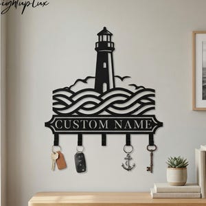 May include: Black metal lighthouse key holder with the text "CUSTOM NAME". The design includes a lighthouse, waves, and hooks for keys. The key holder is mounted on a wall above a wooden surface.