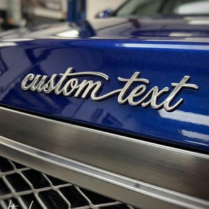 Puede incluir: Primer plano del capó azul de un coche con las palabras "custom text" en escritura plateada. Debajo del texto hay un adorno plateado y una rejilla negra. El logotipo "Lightuplux" está en la esquina inferior izquierda.