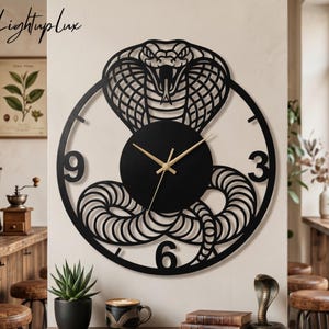 May include: Black metal wall clock featuring a cobra design. The clock has gold-colored hands and the numbers 3, 6, and 9 are visible. The clock is mounted on a light-colored wall. The brand name "Lightuplux" is in the upper left corner.