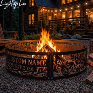 May include: A circular black metal fire pit ring featuring laser-cut butterfly and foliage designs. The metal panels display the text CUSTOM NAME and EST 2026. A bright orange fire burns in the center of the ring, which is placed on gravel.