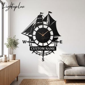 Pode incluir: Um relógio de parede em metal preto com a silhueta de um veleiro integrada num design de rosa dos ventos. O mostrador inclui o texto CUSTOM NAME e os pontos cardeais W, E e S. Ponteiros finos de cor dourada indicam as horas.
