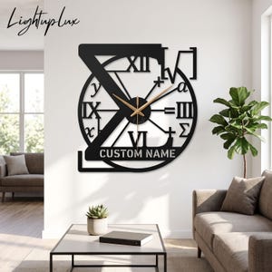 Könnte beinhalten: Eine schwarze Wanduhr mit einem großen Buchstaben Z, der in einen kreisförmigen Rahmen mit römischen Ziffern, mathematischen Symbolen und dem Text CUSTOM NAME integriert ist. Die Uhr hat dünne goldfarbene Zeiger und hängt an einer weißen Wand.