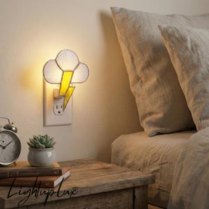 May include: A decorative night light shaped like a cloud with a yellow lightning bolt. The night light is plugged into a white wall outlet. The text "LightupLux" is visible on a wooden nightstand.