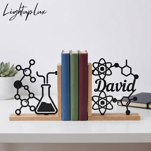 Puede incluir: Un par de sujetalibros de madera con recortes de metal negro. La pieza izquierda muestra un matraz químico y una estructura molecular. La pieza derecha presenta símbolos atómicos y el nombre David en letra cursiva. Tres libros se sostienen.