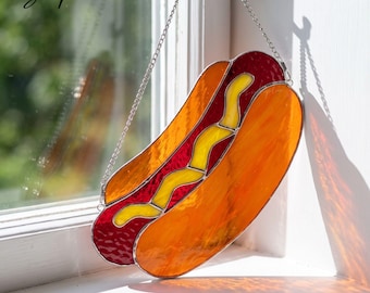 May include: A stained glass suncatcher shaped like a hot dog, featuring orange, red, and yellow glass segments held together by metal solder lines. The piece hangs from a silver-toned metal chain and displays the text Lightuplux in the corner.