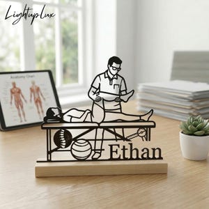 May include: A black metal silhouette desk ornament featuring a physical therapist adjusting a patient on a treatment table. The design includes a spinal column graphic, an exercise ball, and the name Ethan, mounted on a light wood base.