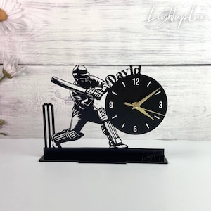 Puede incluir: Reloj negro con temática de cricket que representa a un bateador en acción, con una esfera y el nombre "David" en la parte superior. El reloj tiene manecillas doradas y está montado sobre una base negra. El diseño incluye los postes de cricket.