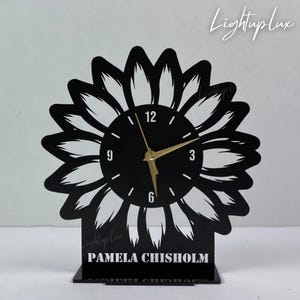 May include: Black sunflower-shaped desk clock with gold hands and a black clock face. The clock has the numbers 12, 3, 6, and 9. The base of the clock has the name "PAMELA CHISHOLM" in white text. The clock is a decorative item.