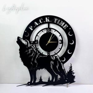 Può includere: Un orologio da parete circolare nero con la sagoma ritagliata di un lupo che ulula alla luna. Il quadrante mostra il testo P.A.C.K. TIME lungo il bordo superiore, con icone a mezzaluna e una scena boschiva alla base. Lancette dorate.