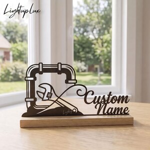 Op de afbeelding: Een donkerbruin lasergesneden bord met een frame van loodgietersbuizen en een afbeelding van een verstelbare moersleutel. De tekst "Custom Name" staat in een cursief lettertype aan de rechterkant. Het bord staat op een lichte houten voet.