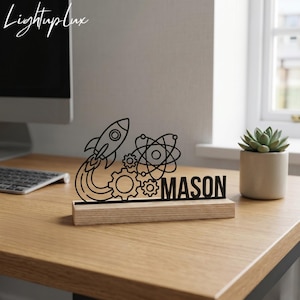 May include: A black metal cutout sign featuring a rocket ship, gears, and an atom symbol, with the name MASON integrated into the design. The cutout is mounted on a rectangular light-colored wooden base, creating a desk ornament.
