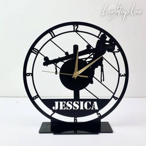 May include: Black laser-cut clock featuring a lineman silhouette, gold clock hands, and the name "JESSICA" at the base. The clock has a circular design with Roman numerals and sits on a black stand. The text "LightupLux" is in the upper right corner.