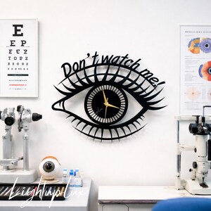 May include: A black wall clock shaped like an eye with long eyelashes and the text Don't watch me! written in a cursive font above the iris. The clock face features a black center with white radial lines and gold-colored hour and minute hands.
