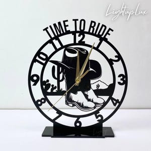 以下が含まれることがあります： カウボーイをテーマにした黒いレーザーカット時計。時計の文字盤には、カウボーイハットとブーツ、砂漠の風景のシルエットが描かれています。「TIME TO RIDE」の文字が上部にあります。 金色の時計の針。