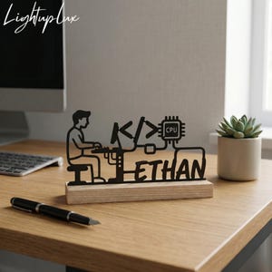 May include: A black metal desk ornament featuring a silhouette of a person sitting at a computer, code brackets, a CPU icon, and the name ETHAN, all mounted on a rectangular light-colored wooden base. The design depicts a programmer at work.