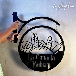 May include: Black metal bakery sign with a circular design featuring bread silhouettes. The sign reads "La Canaria Bakery" in a stylized font. The sign is hanging from a bracket and chain.