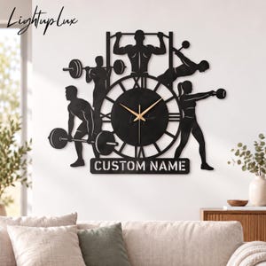 Könnte beinhalten: Eine schwarze Wanduhr mit Silhouetten von Personen bei verschiedenen Fitnessübungen, darunter Gewichtheben und Klimmzüge. Das Zifferblatt hat römische Ziffern und goldfarbene Zeiger. Unter der Uhr befindet sich ein Ausschnitt mit dem Text CUSTOM NAME.