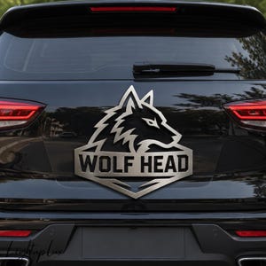Puede incluir: Un emblema de coche de color plata metálica que presenta una silueta estilizada de cabeza de lobo sobre una pancarta que contiene el texto WOLF HEAD en letras mayúsculas negritas. El emblema está montado en el portón trasero de un vehículo oscuro.