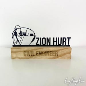以下が含まれることがあります： ヘルメットをかぶり設計図を持つエンジニアのシルエットとZION HURTの文字が描かれた黒い金属製のデスクサイン。金属部分はCIVIL ENGINEERと刻印された明るい色の木製ベースに取り付けられています。