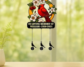 Könnte beinhalten: Ein kreisförmiges Hängeornament mit einem roten Kardinal, der auf einem Ast mit weißen Blüten und roten Kirschen sitzt. Unter dem Kreis befindet sich eine schwarze Plakette mit dem Text IN LOVING MEMORY OF WILLIAM - 1950-2025 und drei hängenden Vogelanhängern.