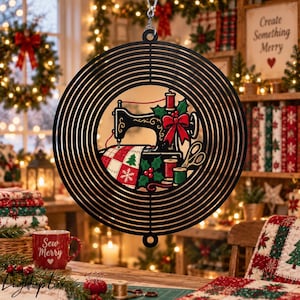 May include: A circular black metal wind spinner featuring a central graphic of a vintage sewing machine decorated with a red bow, holly leaves, and berries. The design includes a spool of thread, scissors, and a fabric piece with a snowflake pattern.
