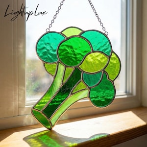 May include: A stained glass broccoli design in shades of green, hanging from a silver chain. The artwork features a detailed depiction of the vegetable, with varying shades of green glass. The piece is displayed near a window, catching the sunlight.