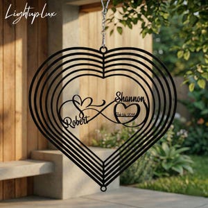 May include: Black metal heart-shaped wind spinner with the names "Robert" and "Shannon" and the date "Feb 14 2016" cut out. The spinner has a layered design and is suspended by a chain.