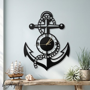 May include: A black wall clock shaped like a nautical anchor with a rope design wrapped around the center. The clock face is circular with black numbers and thin gold hands. The design is flat and cut out from a solid material, mounted on a wall.