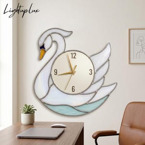 Puede incluir: Un reloj de pared con forma de cisne con un diseño de vidrieras blancas y azules. La esfera del reloj es redonda con manecillas y números dorados. El texto "Lightuplux" es visible en la esquina superior izquierda.