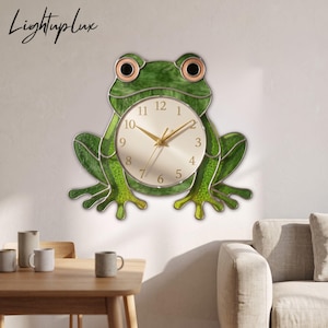 May include: A frog-shaped wall clock featuring green stained glass panels with dark veining, large round eyes with peach-colored centers, and a central circular clock face with gold-toned numbers and hands. The text LightupLux is visible.