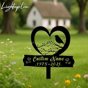 May include: Black metal garden stake with a heart-shaped design featuring a bird on a branch. The stake includes the text "Custom Name" and the years 1975-2021. The stake is placed in a grassy area with a house in the background.