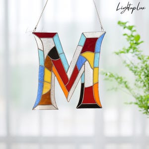 May include: A colourful stained glass letter 'M' with a silver chain for hanging. The letter is composed of various coloured glass pieces, including red, blue, yellow, and brown. The word "Lightuplux" is written in cursive.