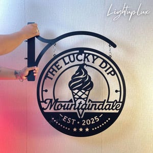 Puede incluir: Letrero de metal negro para una heladería, con el texto "The Lucky Dip" y "Mountaindale EST. 2025". El letrero tiene un diseño circular con un gráfico de cono de helado y está sujeto por un soporte de metal negro.