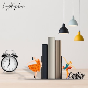 May include: A black metal bookend set featuring two dancing figures, one in an orange floral dress and one in a teal leotard, holding three books. The set includes a black analog alarm clock and the text LightupLux and Custom name.