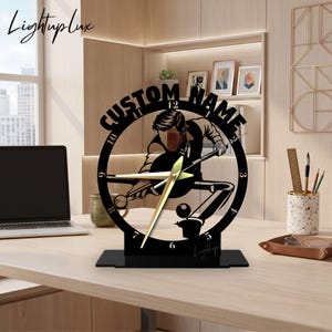 May include: A black circular desk clock featuring a cutout silhouette of a person playing billiards. The clock face displays the text CUSTOM NAME at the top, numbers 1 through 12, and gold-colored hands. It stands on a rectangular black base.