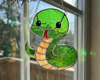May include: A stained glass suncatcher shaped like a green snake with a light yellow underbelly and a small red tongue. The snake has two large black circular eyes and is suspended by a silver metal chain against a window pane.