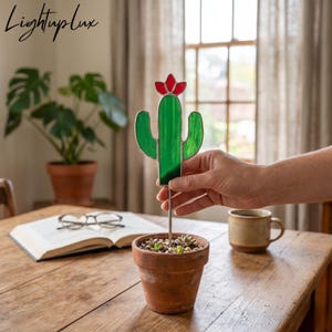 Puede incluir: Un adorno de cactus de vidrieras con cuerpo verde y una flor roja en la parte superior, montado sobre una fina varilla de metal. El cactus está colocado dentro de una pequeña maceta de terracota llena de tierra y pequeñas suculentas, con la etiqueta LightupLux.