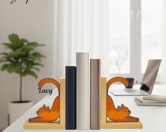 Puede incluir: Un par de sujetalibros con forma de gato naranja y acabado liso. Cada sujetalibros presenta un gato estilizado en pose de estiramiento. El sujetalibros izquierdo tiene el nombre Lucy impreso en letra cursiva negra.