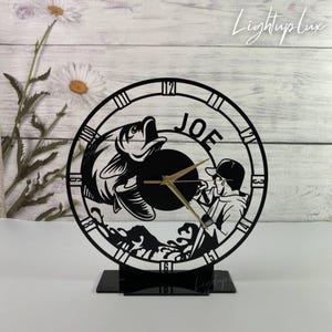 May include: Black laser-cut clock featuring a fisherman reeling in a fish, with the name "JOE" above the clock face. The clock has gold-colored hands and is mounted on a black base. The clock is set against a white wooden background.