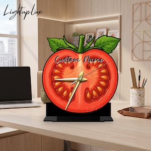 May include: A tomato-shaped desk clock featuring a cross-section illustration of a red tomato with green leaves at the top. The clock face displays the text Custom Name in black script and includes gold-colored hour, minute, and second hands.