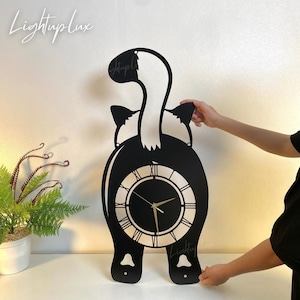 May include: A black and white skunk-shaped wall clock with Roman numerals. The clock has a circular face and is held by a person. The clock has the text "LightupLux" in the upper left and lower right corners.
