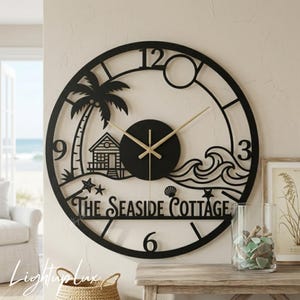 May include: A circular black metal wall clock featuring a cutout design of a palm tree, a small cottage, ocean waves, and seashells. The text THE SEASIDE COTTAGE is integrated into the lower half. It has gold-coloured hands and numbers 3, 6, 9, and 12.