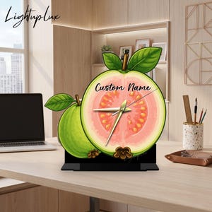 May include: A guava-shaped desk clock featuring a cross-section of a pink guava with seeds and green leaves. The clock face displays the text Custom Name in black script, gold-coloured clock hands, and is mounted on a black rectangular base.