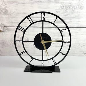 May include: A black circular skeleton clock with a stand, featuring large cut-out numerals from 1 to 12 arranged around the perimeter. The center has a solid black circular dial with gold-colored hour and minute hands. The text Lightuplux is visible.