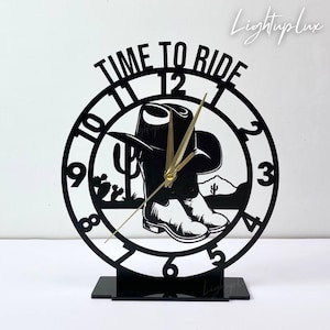 以下が含まれることがあります： カウボーイをテーマにした黒いレーザーカットの卓上時計。時計には、カウボーイハットとブーツ、砂漠の風景、そして「TIME TO RIDE」の文字が描かれています。時計の針は金色です。