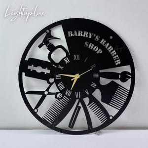 May include: Black barber shop clock with gold hands and Roman numerals. The clock features barber tools like scissors, combs, and a straight razor. The text "BARRY'S BARBER SHOP" is displayed on the clock face. The clock is a decorative item.