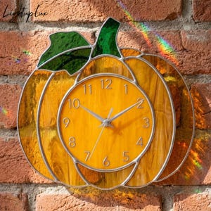 May include: A stained glass wall clock shaped like a pumpkin with a yellow face, silver hands, and black numerals. The pumpkin body is made of amber-colored glass with a green glass leaf at the top. The text Lightup lux is visible.