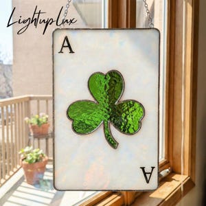 May include: A rectangular stained glass suncatcher shaped like an ace of spades playing card, featuring an iridescent white background and a textured green shamrock in the center. The letter A is printed in the top left and bottom right corners.