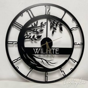 May include: Black metal wall clock with a tree design. The clock features Roman numerals and the name "WILHITE" in a horizontal band. The clock hands are gold-colored. The clock is a decorative item for home decor.